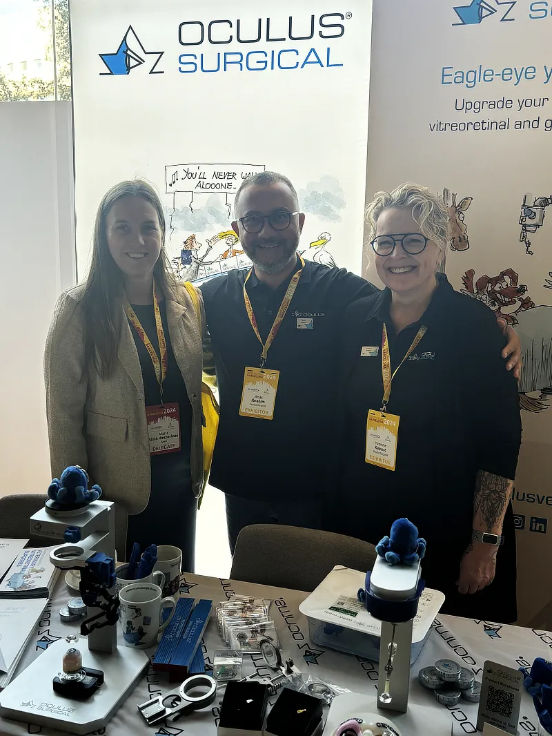 La Dra. María Simó posa con dos miembros del equipo de Oculus Surgical en su stand del congreso ESVO 2024. Se observan dispositivos quirúrgicos en la mesa del stand, utilizados para cirugía de retina.