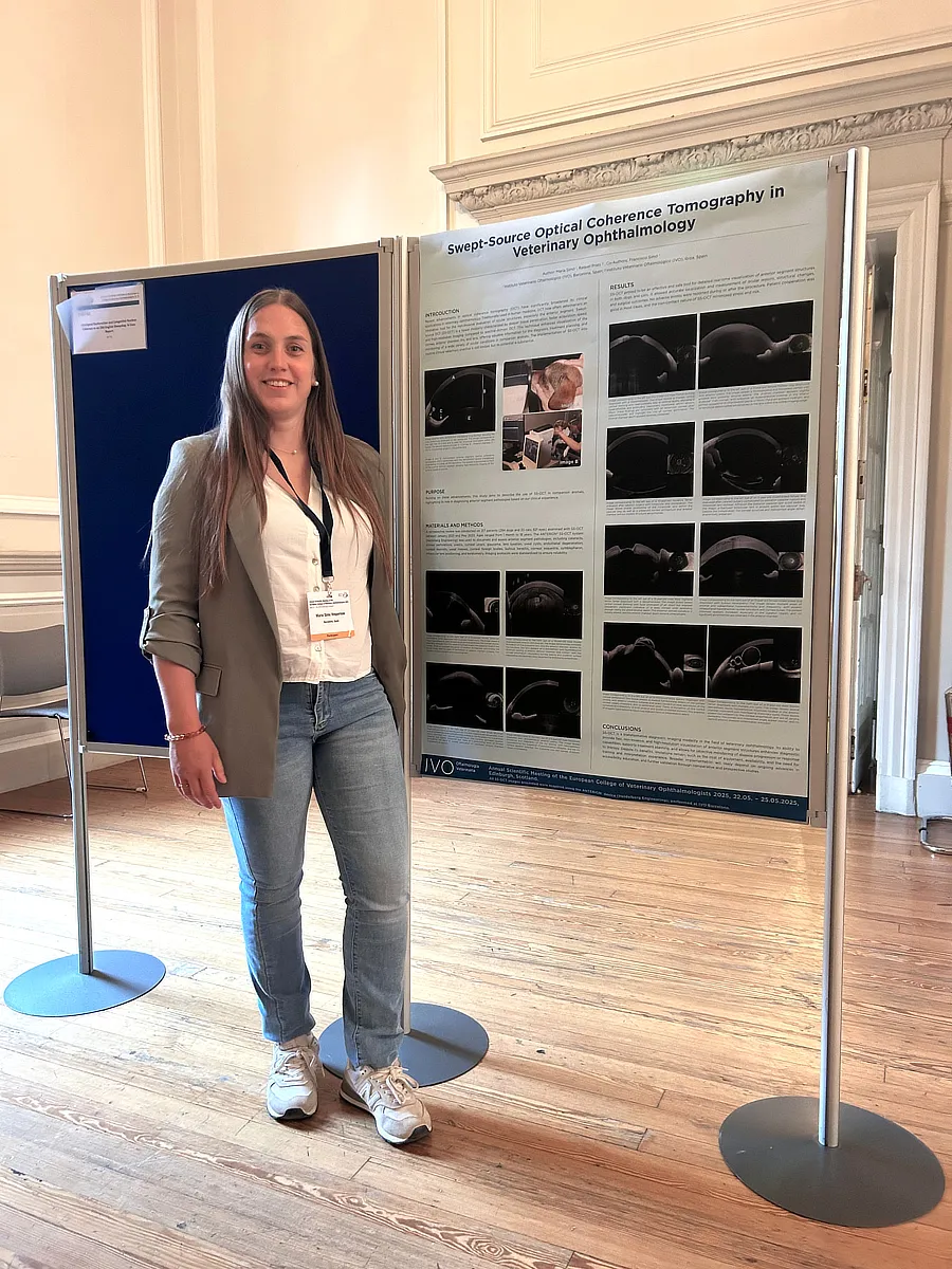 Dra. María Simó posando de pie junto a un póster científico sobre SS-OCT en animales, montado sobre paneles verticales en una sala iluminada.